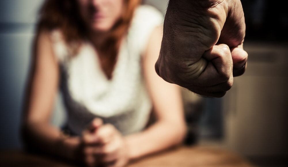 Young woman sat at a table in fear of domestic abuse, with a clenched fist in the foreground