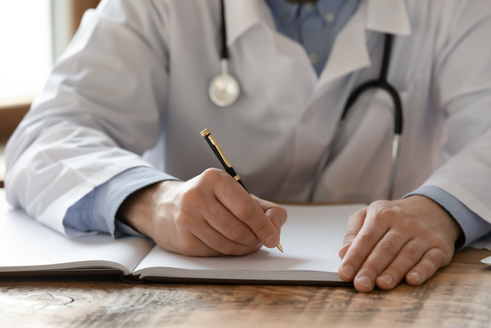 Close-up of GP in white lab coat filling in a form at his desk