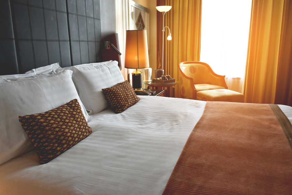 Photograph of the inside of a hotel room with a double bed with cream and brown duvet, chair, lamp and bedside table