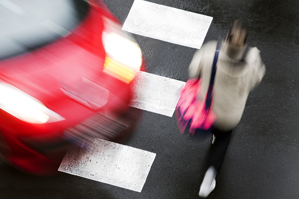 Photograph of woman pedestrian crossing the road about to be hit by a red car