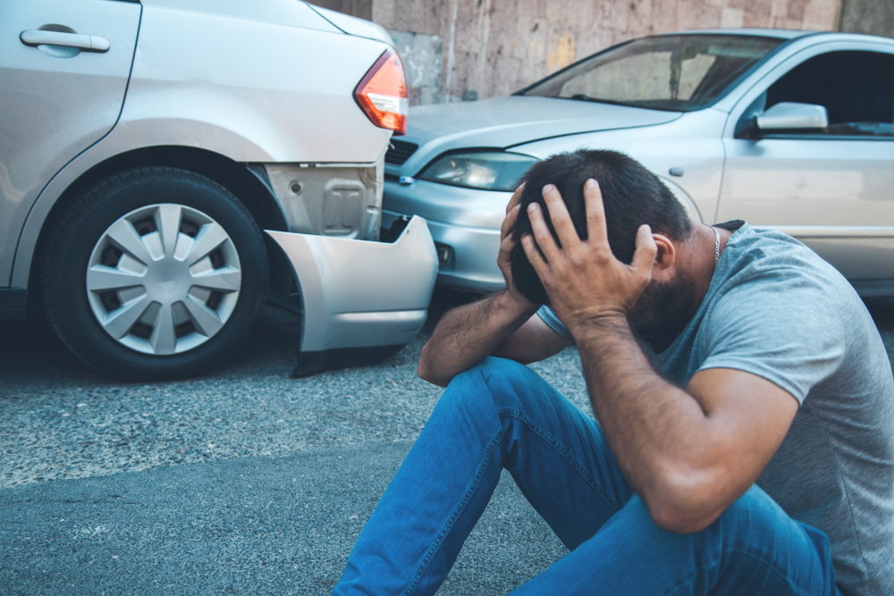 Photograph of man at down on the road with his head in his hands after a car crash accident in the background