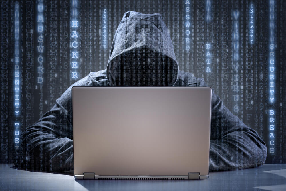 Photograph of ominous hooded figure working at a laptop to steal data, with broken code as a background