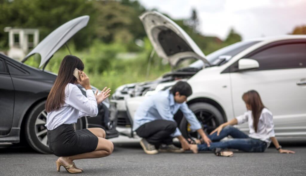 Woman on the phone reporting a road traffic accident to insurance company, with man helping injured woman's leg next to two smashed cars in background