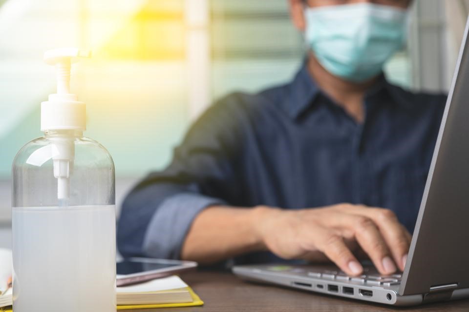 Employee wearing PPE in an office with his laptop and bottle of anti-bacterial gel on his desk