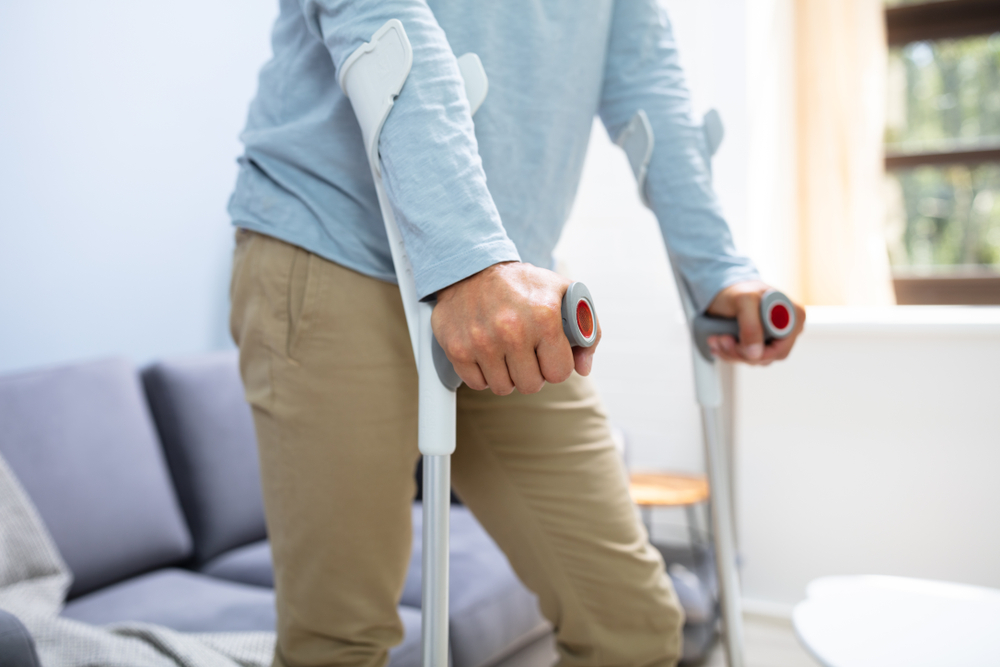 Photograph of man in a blue shirt and brown trousers using crutches to walk around house after work injury