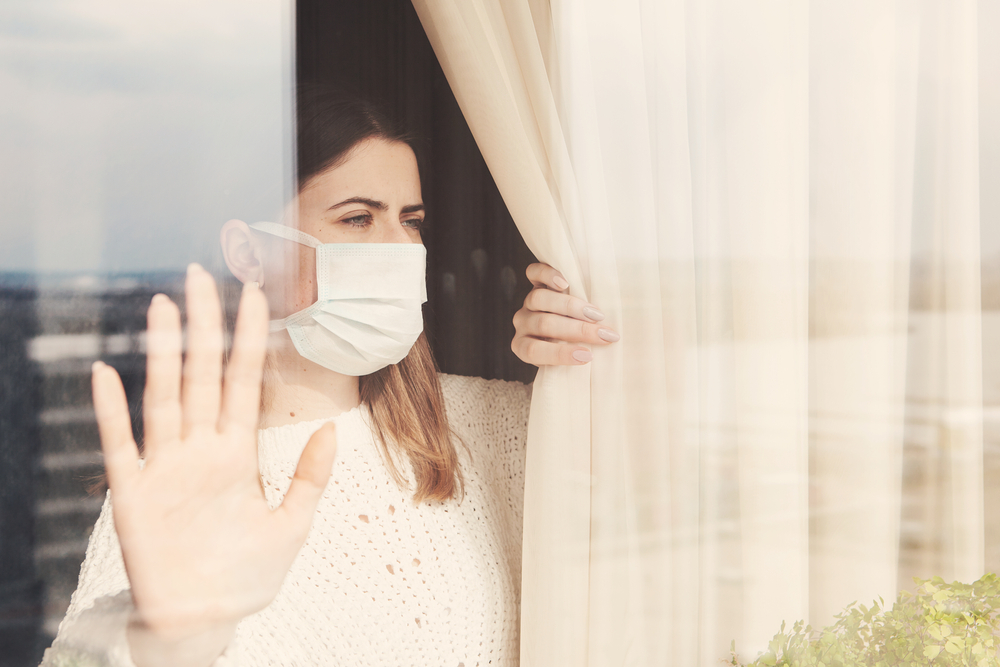 Photograph of a student in a COVID mask looking outside of her window during lockdown