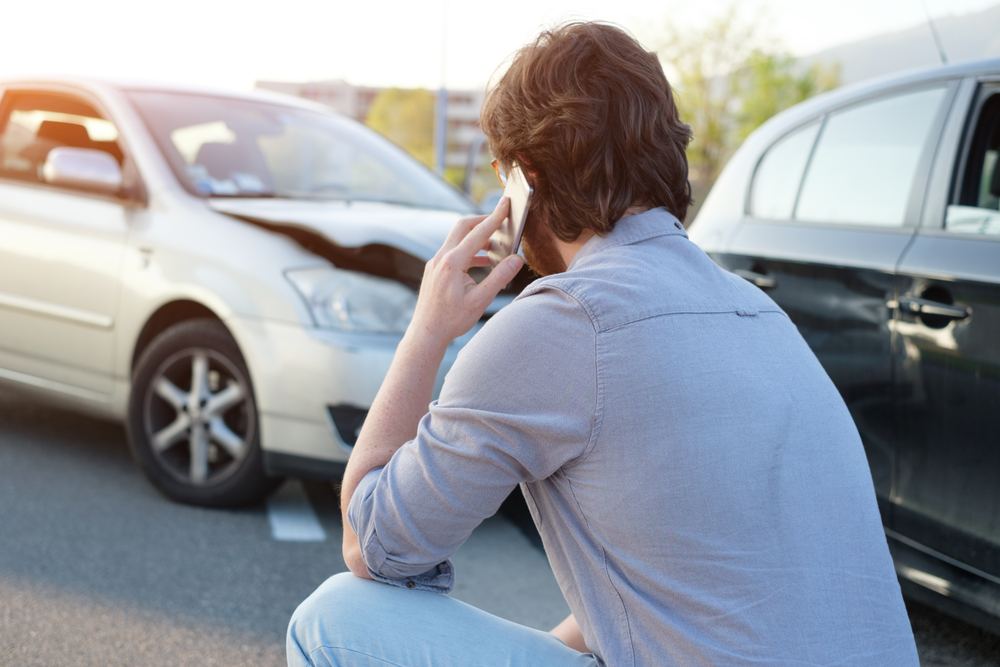 Man crouched on the ground calling for help on his phone after a road traffic accident between a white and black car