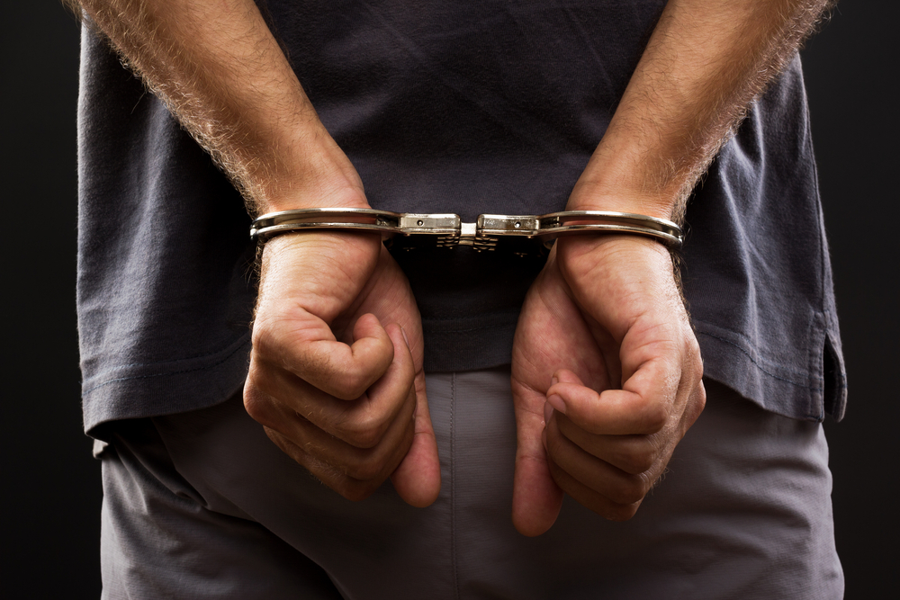 Photograph of a man with his hands cuffed behind his back against a dark background