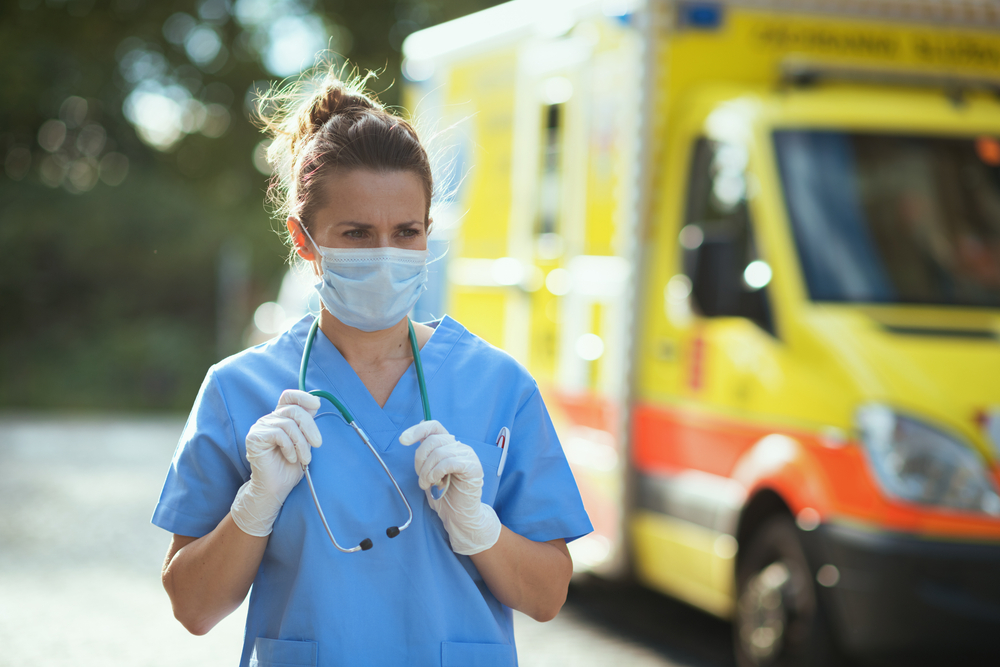 Photograph of ambulance worker in scrubs and surgical mask next to an ambulance vehicle 