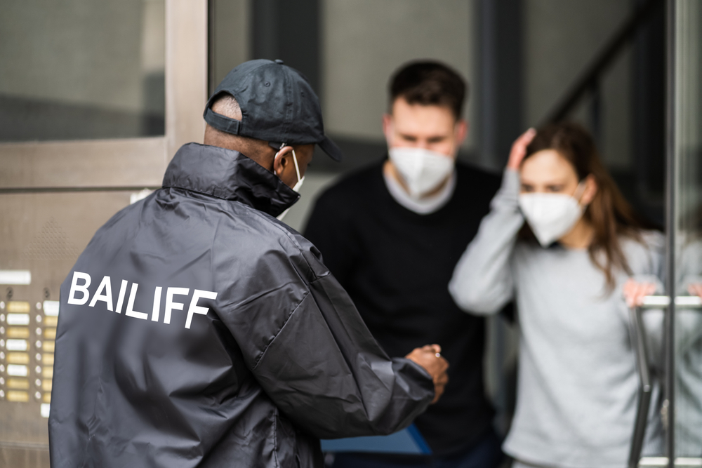 Photograph of bailiff in black jacket next to a woman and man in their house wearing surgical masks having their possessions reclaimed