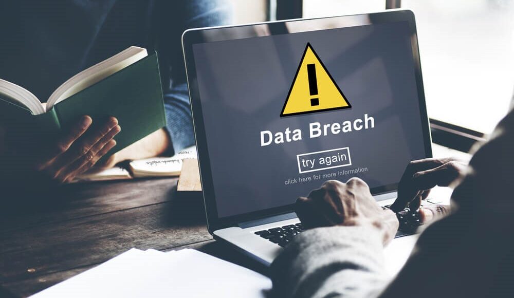 A colour image of two people sat at a table across from each other, the one closest to the camera is typing on a laptop which displays a yellow caution symbol above the words 'Data breach' and a button which says try again, click here for more information