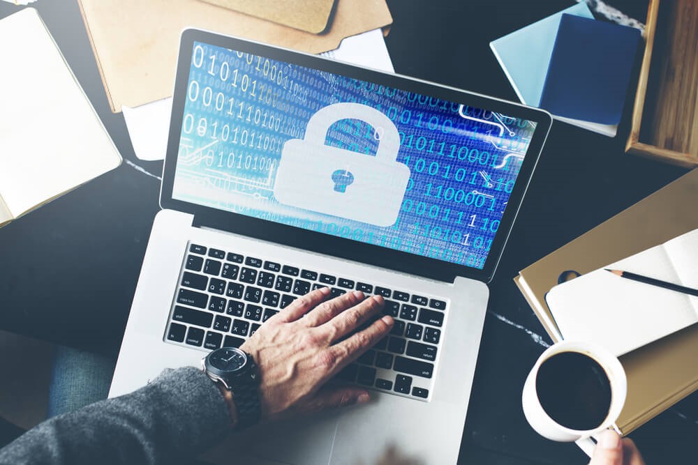 Colour picture of a person typing on a silver laptop with a screen displaying a blue background covered in white coding and a large white padlock in the middle of the screen