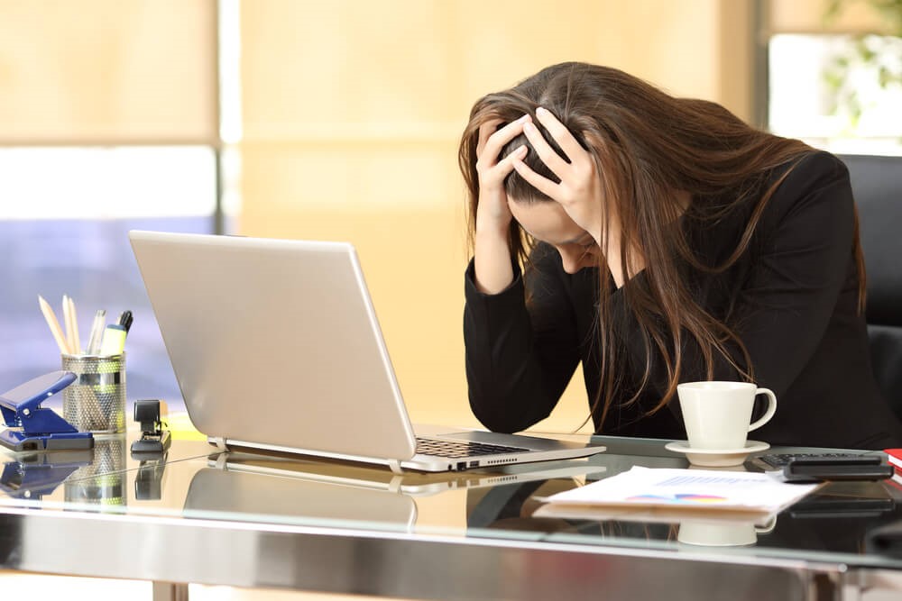 A colour picture of an emotionally distressed woman sat at a desk with her head in her hands