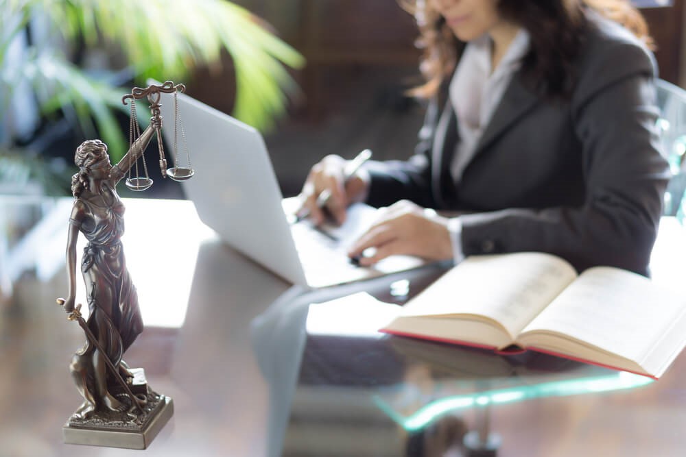 Colour image of a solicitor sat at a desk typing on a laptop with a book to her side and in the foreground is a silver statue of lady justice - a woman holding scales 