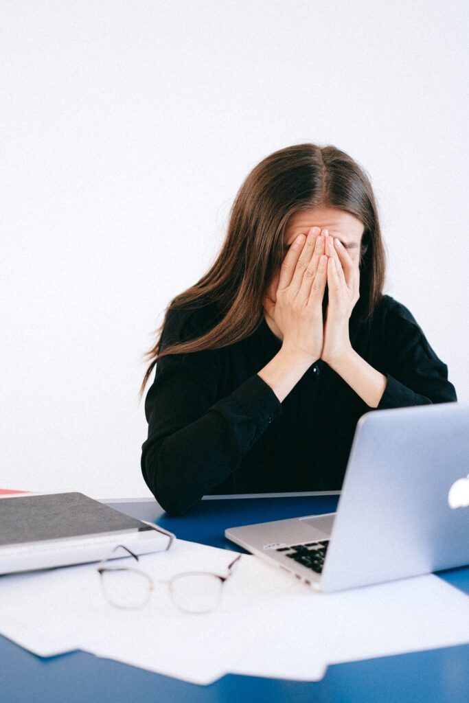 A woman with long brown hair in a black jumper, sat in front of an open laptop with her face in her hands.