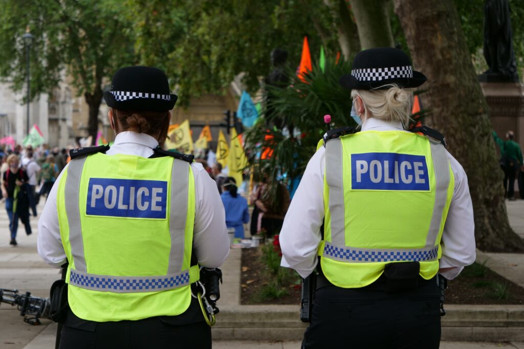 Two police officers in high-visibility vests walking away from the camera toward a busy shopping area.