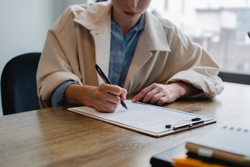 A woman holding a pen above a clipboard while sitting at a wooden desk.