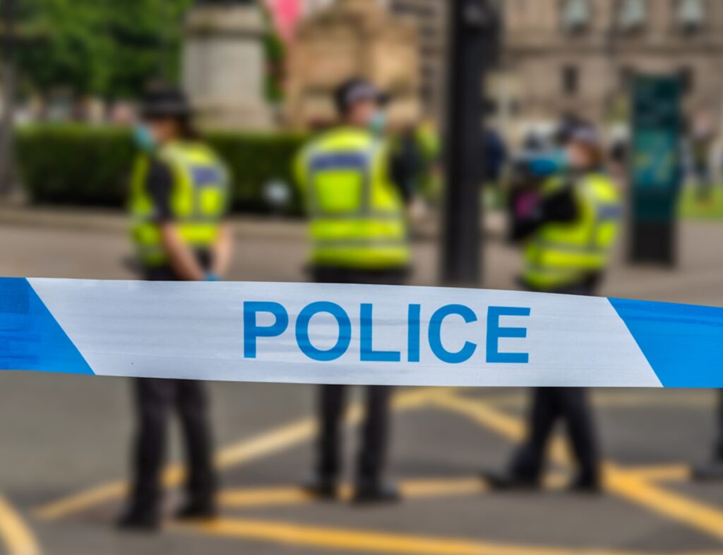 Blue and white police tape in the foreground with three police officers standing on a street in the background.