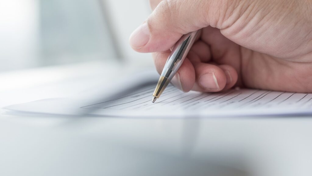 A hand holding a silver and gold pen signing a document.