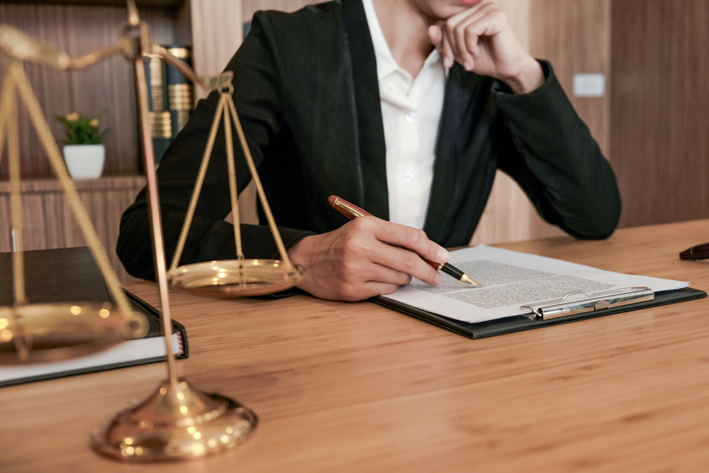 An image of a solicitor sat at a wooden desk reading a document whilst holding a pen. In the foreground is a pair of scales - legal concept.
