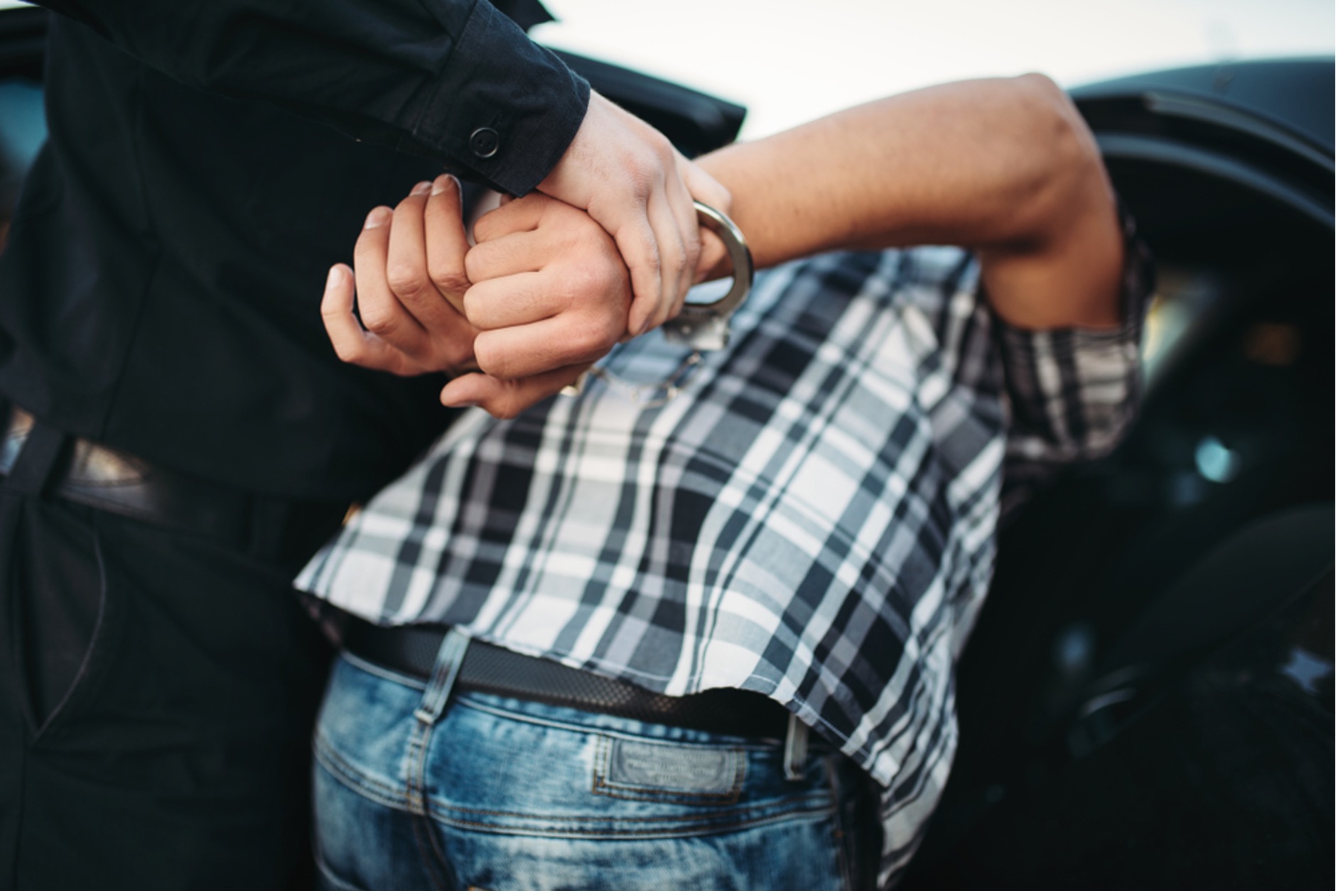Colour image of a man wearing handcuffs being arrested and placed in the back of a police car by a police officer