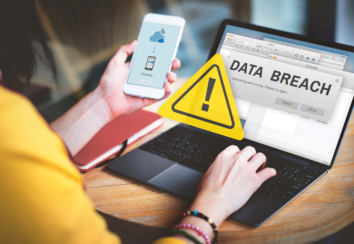 Data breach concept image, a woman in a yellow top holding a mobile phone in front of a laptop with a notification saying 'DATA BREACH' with a warning symbol