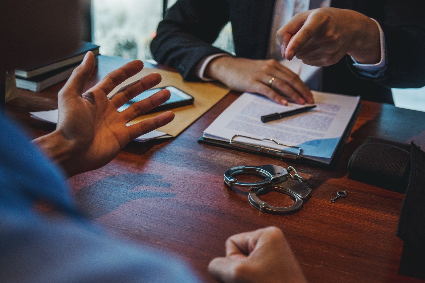 Solicitor concept image, person at at a desk talking to a solicitor, the solicitor has papers on a clipboard in front of them and is pointing at the person sat opposite them. On the table is a pair of handcuffs