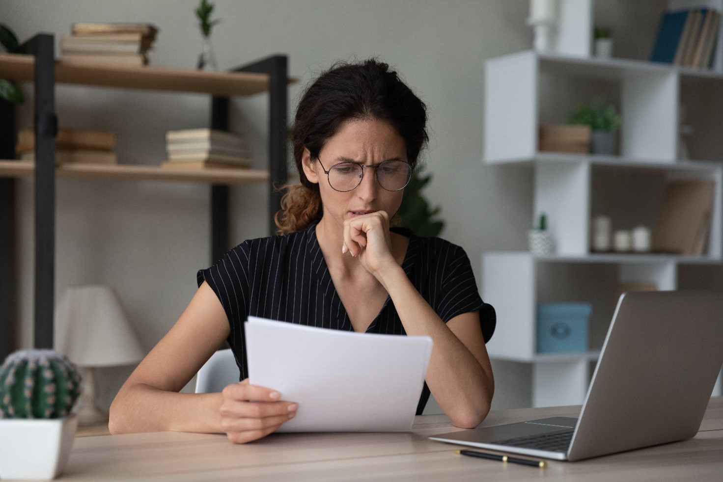 Confused and distressed looking woman reading something of a piece of paper sat at a desk in front of a laptop