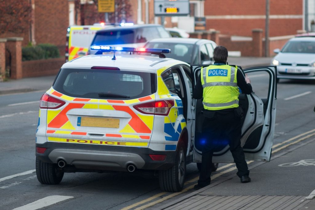 Image of the back of a police car with its lights flashing and a police officer stood at the open back door of the police car