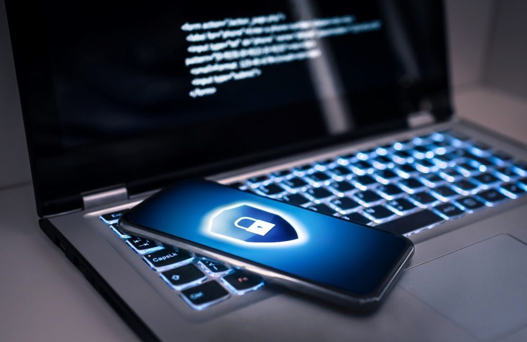 Image of a laptop with a phone lying on the keyboard, the screen shows a data security symbol - a shield with a padlock inside