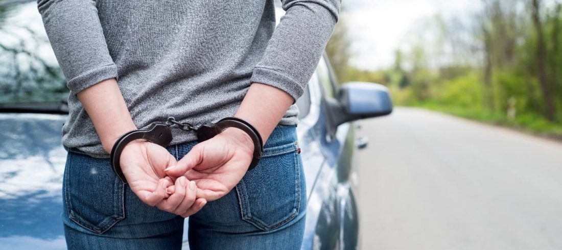 Image of a handcuffed woman with her hands behind her back stood on a road in front of a blue car