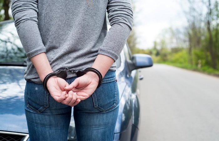 Image of a handcuffed woman with her hands behind her back stood on a road in front of a blue car