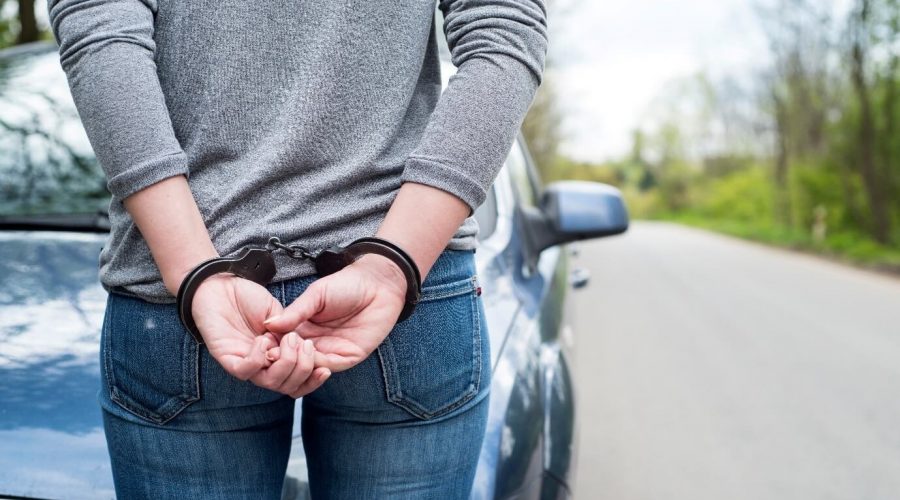 Image of a handcuffed woman with her hands behind her back stood on a road in front of a blue car