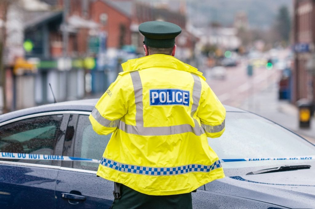 A police officer wearing a high-visibility jacket stood in front of police tape and a car on the street