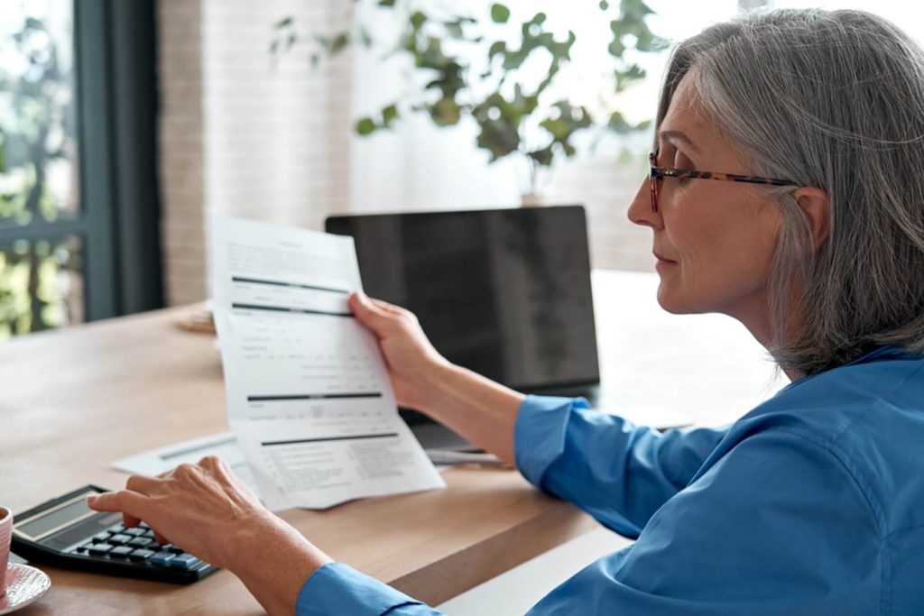 An retired woman reading about her pension from a piece of paper, calculating something on a calculator. She's sat at a table with a laptop to the side of her and a coffee. 