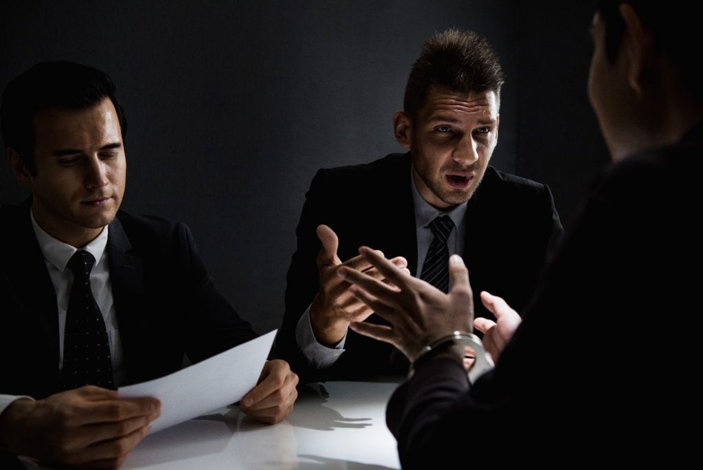 Two police officers in suits sat interviewing a suspect who is in handcuffs.