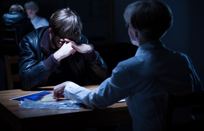 Image of a police interview. A woman police officer is interviewing a male who is holding his head in his hands. The officer is pointing to a picture on the table.