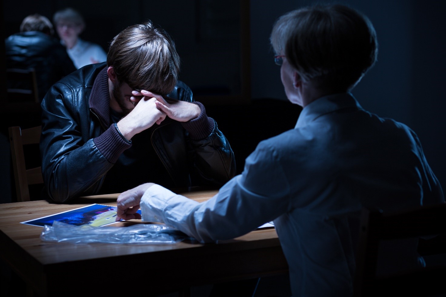 Image of a police interview. A woman police officer is interviewing a male who is holding his head in his hands. The officer is pointing to a picture on the table.