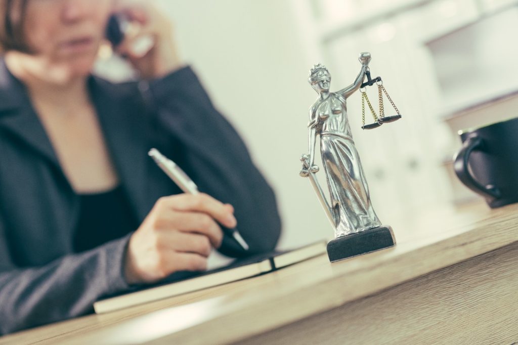 An image of lady justice statue on a desk in from of a solicitor who is in the background on a mobile phone and writing in a notebook,