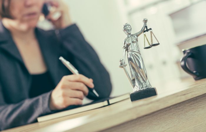 An image of lady justice statue on a desk in from of a solicitor who is in the background on a mobile phone and writing in a notebook,