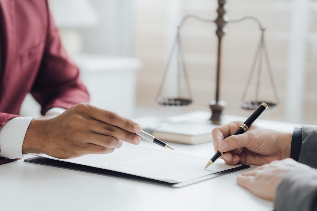 A solicitor and client sat face-to-face, the client is signing a document and the solicitor is pointing to a section of it with their pen.