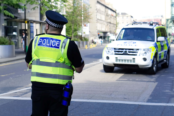 Photograph of a police officer stood in the street in front of a white police vehicle 