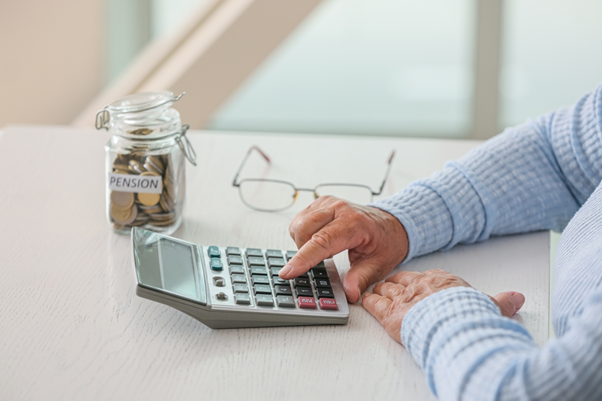 A picture of an elderly person working out an equation on a calculator sat behind a table with a pot full of coins that says 'pension' on it next to them. 