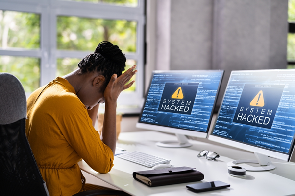 A woman sat behind a desk looking stressed, with the palms of her hands pressed to her head. In front of her on the desk is a two monitors with warning messages saying "System hacked" on each. 