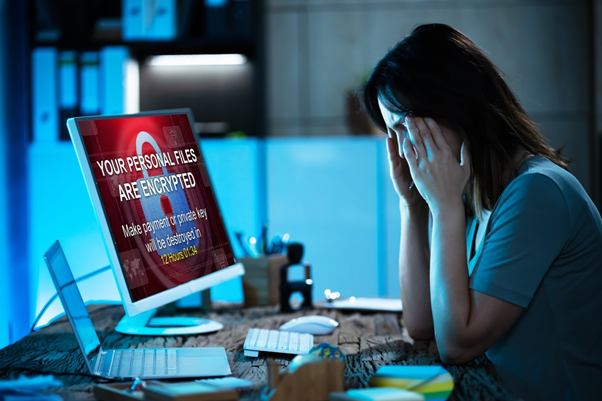 Picture of a woman sat at a desk looking distressed holding her hands to her temples. In front of her on the desk is a computer which has a warning message on the screen stating "Your personal files are encrypted. Make a payment or private key will be destroyed in 12 hours".
