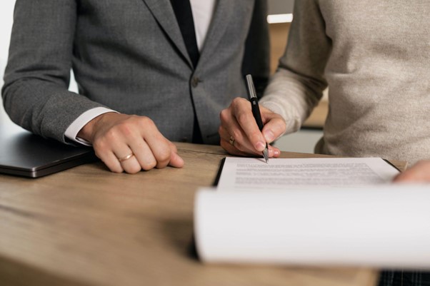 Two people stood at a table reading a document, one has a pen in hand and is signing the page.