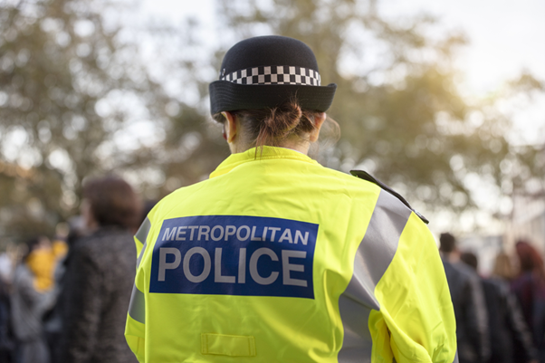 A picture of the back of a female Metropolitan police officer wearing a black police hat and a high-visibility neon green jacket with "Metropolitan Police" written on the back of it.