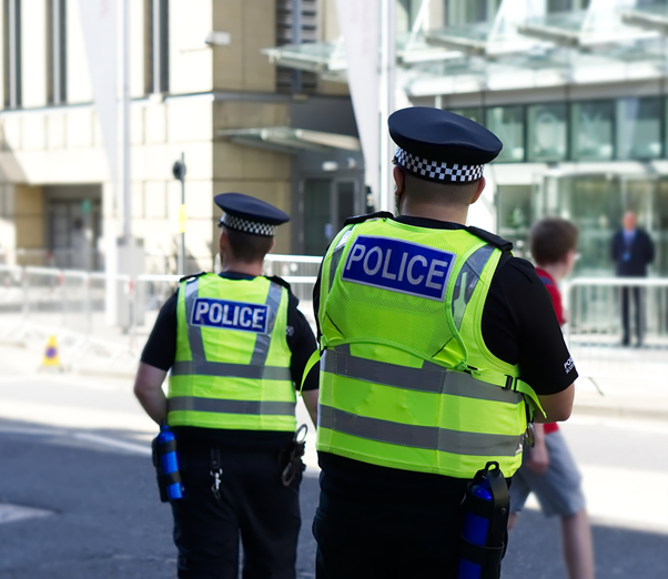 A photograph of two UK police officers stood on the street outside a large building. Both are officers are wearing high visibility police vests. 