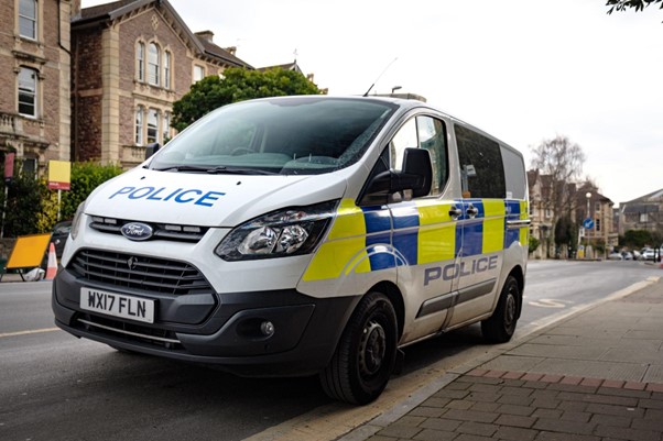 A photograph of a white police van with blue and fluorescent yellow squares down the side parked up on a street
