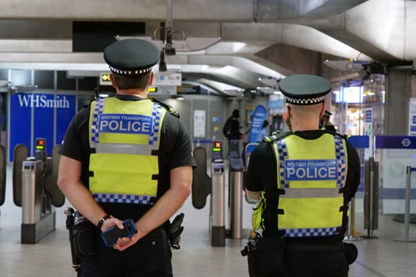A photograph of two male police officers stood in a train station with their backs to the camera. They are both wearing high-visibility vest with the text "British transport police" written on the back. 